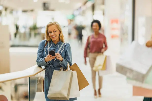 Happy female shopaholic using phone on social media to search discount codes for sales while shopping at the city mall.