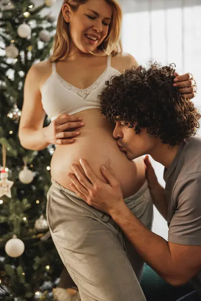 Genç adam hamile karısının karnını öpüyor ve eğleniyor. Mutlu bir yeni yılı evde kutluyor..