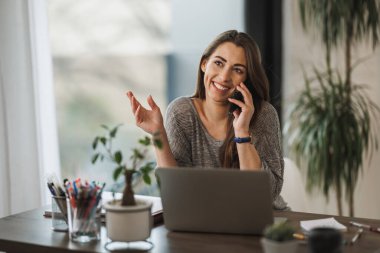 Evde dizüstü bilgisayarla çalışırken cep telefonu kullanan mutlu genç bir kadın..