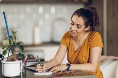Genç bayan tasarımcı, stüdyosunda bilgisayar üzerinde çalışırken notlar alıyor..