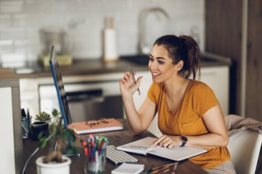 Genç bayan tasarımcı ev ofisinden bilgisayar üzerinde çalışırken notlar alıyor..