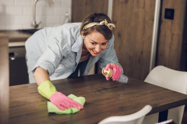 Evde mutfak masasını temizlerken gülümseyen mutlu kadın. Koruyucu eldiven takıyor ve sprey kullanıyor..