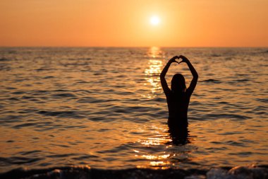 Tatilde deniz kenarında gün batımının keyfini çıkarırken kendini özgür hisseden genç bir kadının silueti. Elleri okyanusun üzerinde kalbi şekillendiriyor..