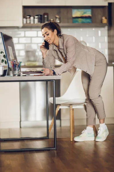 Young woman designer talking on a smartphone while working on computer from her home office.