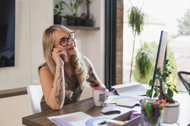 Genç, gülümseyen bir kadın evinde rahat ederken cep telefonu kullanıyor ve kahve içiyor..