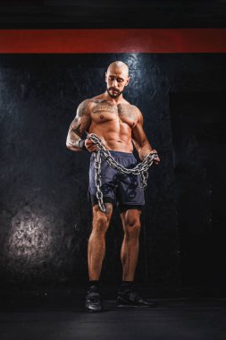 Genç bir fitness adamı salonda duruyor ve ağır bir zincirle antrenman yapmaya hazırlanıyor..