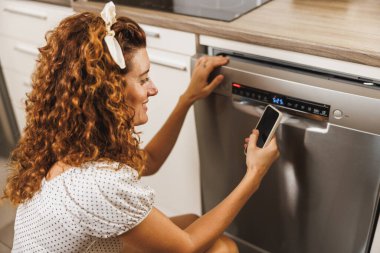 Evdeki mutfağında bulaşık makinesini kontrol etmek ve açmak için akıllı telefon kullanan güzel bir kadın..
