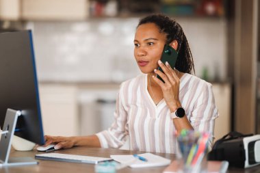 Ofisinden bilgisayar başında çalışan Afrikalı iş kadını akıllı telefondan konuşuyor..