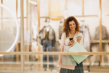 Alışveriş merkezinde alışveriş yaparken akıllı telefonunu kullanarak kart ödemesi yapan genç bir kadının fotoğrafı..
