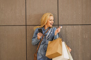 Çok mutlu bir kadının alışveriş torbalarını tutarken akıllı telefonuyla duvar arkasından kart ödemeleri yaparken görüntüsü..