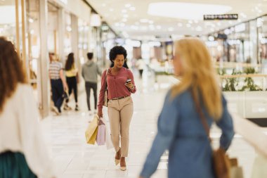 Gülümseyen siyah kadın akıllı telefonunu alışveriş merkezinde yürürken ve çanta taşırken kullanıyor..