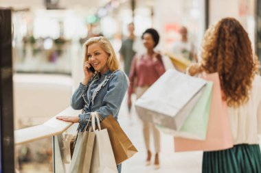 Alışveriş merkezinde alışveriş yaparken çantalarını taşırken telefonu kullanan mutlu bir kadın..