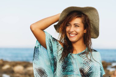 Güneşli bir günde güzel genç bir kadın sahilde dinleniyor. Poz veriyor ve yüzünde tebessümle uzaklara bakıyor.. 