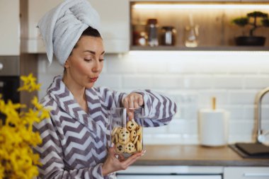 Bornozlu genç bir kadın elinde kurabiye kavanozuyla sabah mutfağında eğleniyor..