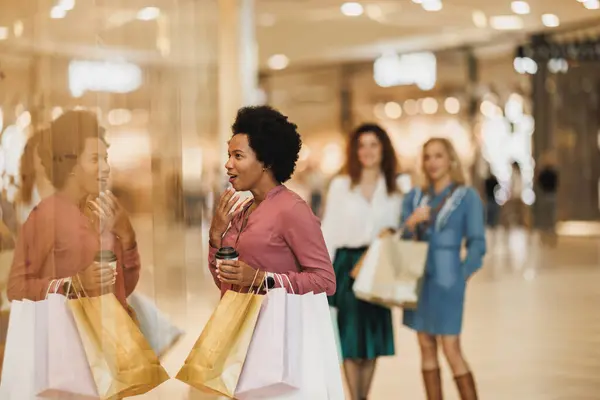 A happy Black woman with bags looks at something in the window of a boutique while enjoying a shopping spree in the city mall.
