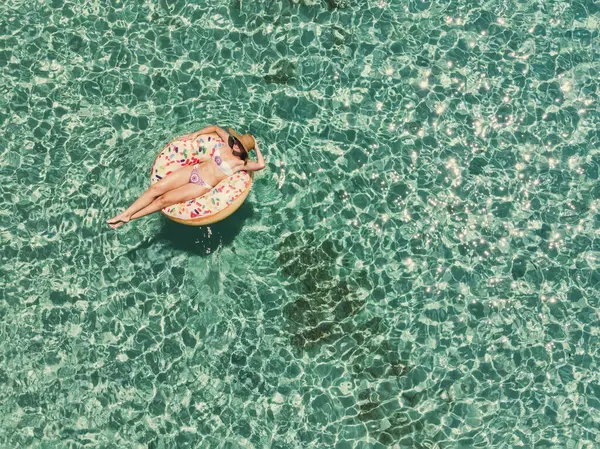 在空中俯瞰着一个美丽的女人 她正在透明的碧绿大海中 在充气的大甜甜圈上游泳 无人机俯瞰 免版税图库照片