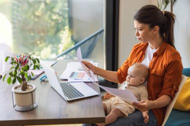 Evde çalışırken ve bebeğini kucağında tutarken endişeli görünen bir kadının fotoğrafı..