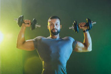 Dambılla ağır antrenman yapan kaslı bir vücut geliştiriciyi vurmuşlar. O, omuzlarını ağır ağırlıkla şişiriyor..