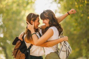 Sırtlarında sırt çantaları olan iki güzel kız arkadaş güneşli caddede yürüyor ve sarılıyorlar..