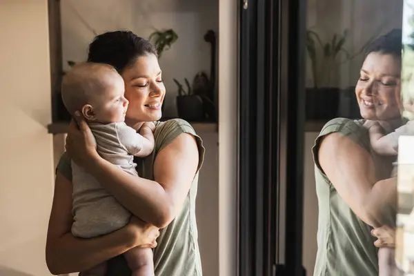 愛する母親のショット 愛らしい赤ん坊を抱えながら 自宅の窓の近くで一日を楽しみながら ストックフォト