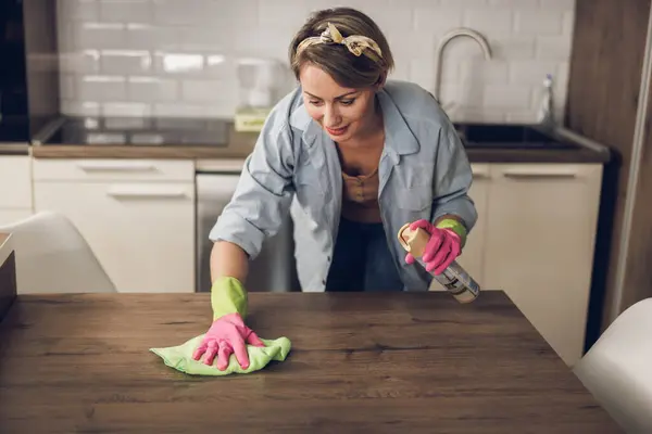 ハッピーな女性が家を清潔に保ち 自宅のキッチンテーブルを拭き取ります 彼女は保護手袋を着てスプレーボトルを使用しています ロイヤリティフリーのストック画像