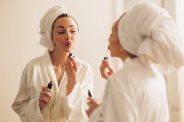 Sabahın keyfini çıkarırken bornozlu çekici bir kadın büyük aynanın önüne dudak parlatıcısı sürüyor..