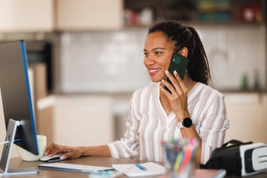 Afrikalı iş kadını ev ofisinden bilgisayarla çalışırken akıllı telefondan konuşuyor..