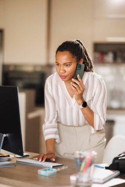 Afrikalı iş kadını düşünceli görünüyor ve evinde bilgisayar başında çalışırken akıllı bir telefonla konuşuyor..