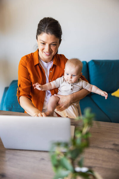 Снимок привлекательной молодой женщины, работающей дома с ребенком на коленях.