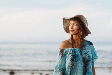 Güneşli bir günde güzel genç bir kadın sahilde dinleniyor. Poz veriyor ve yüzünde tebessümle uzaklara bakıyor.. 
