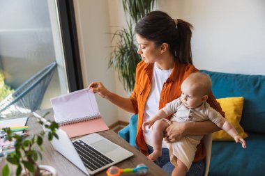 Bebeği kucağında otururken evde çalışan meşgul bir anne..