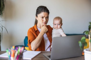 Evde dizüstü bilgisayarla çalışırken bebeğine bakan genç bir annenin fotoğrafı..