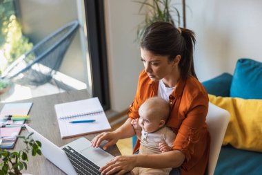 Evde dizüstü bilgisayar başında bebeğini kucağında oturtmakla meşgul bir anne..