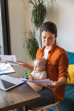 Evde çalışırken stresli görünen ve bebeğini kucağında tutan bir kadın girişimcinin fotoğrafı..
