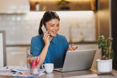 Genç bayan girişimci ev ofisinden bilgisayarla çalışırken akıllı bir telefonla konuşuyor..