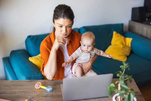 Съемка молодой матери, заботящейся о своей малышке во время работы на ноутбуке из дома.