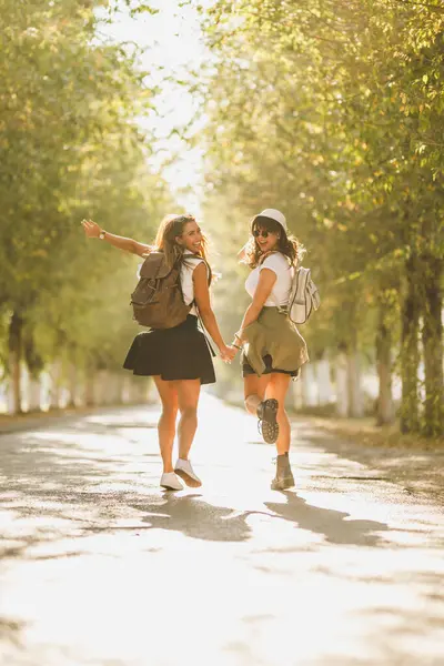 stock image Rear view of a two beautiful cheerful young women with backpacks on their back are having fun while walking along the sunny avenue.