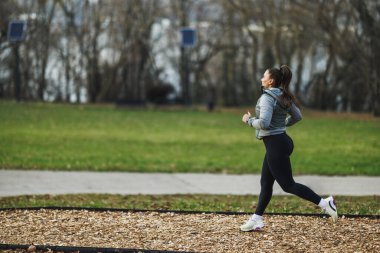 Güneşli bir sonbahar gününde, arka planda uzun ağaçlar olan güzel yapraklı bir parkta neşeyle koşan bir kadın..