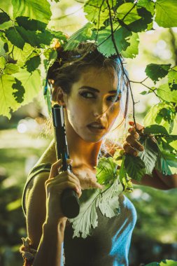 Elinde silah olan odaklanmış bir kadın yeşil ormandaki yapraklar arasında dikkatle dikizliyor..