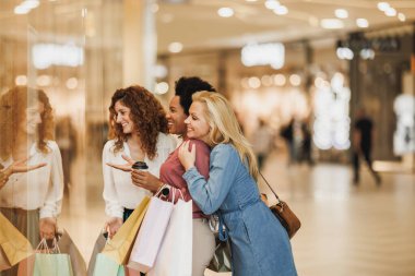 Şehirdeki alışveriş merkezinde alışveriş yaparken butik vitrinine bakan üç gülümseyen kız arkadaşın fotoğrafı.