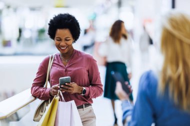 Çok mutlu bir siyahi kadının cep telefonuyla mesajlaşırken ve alışveriş merkezinde alışveriş torbaları tutarken gülümsemesi..