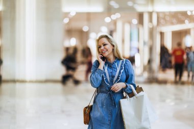 Meşgul bir alışveriş merkezinde çanta taşırken cep telefonuyla konuşan nişanlı bir kadın..