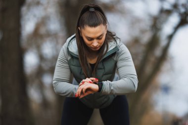 Odaklanmış genç bir kadın akıllı hareketlerini kontrol etmek için durur ve parkta egzersiz yaparken ilerlemesini izler..