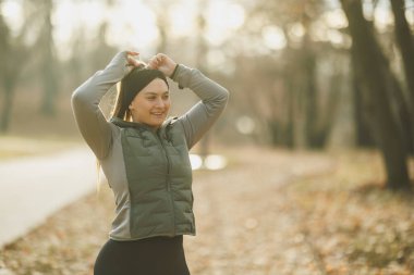 Genç, gülümseyen bir kadın, sabah egzersizine devam etmek için saçlarını sakin, puslu bir park ortamında bağlamaya hazırlanıyor..