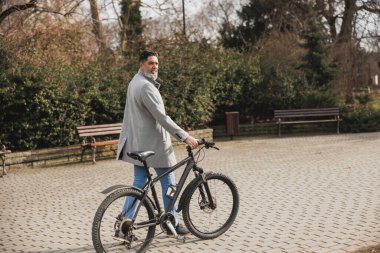 Sıradan bir iş adamı, bisikletiyle tuğla bir yolda yürüyor ve şehir parkında güneşli bir günün tadını çıkarıyor..