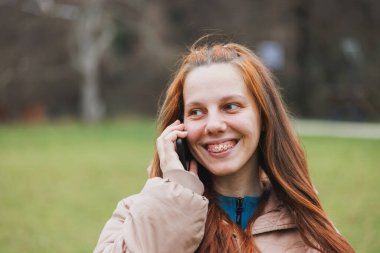 Bir şehir parkında ağaçlar ve yeşilliklerle çevriliyken telleri olan bir kadın cep telefonuyla konuşuyor..