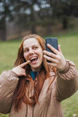 Gülümseyen bir kadın, parkta dikilirken bir fotoğraf çekmek için cep telefonunu kaldırırken görülüyor..