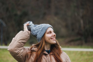 Kahverengi ceketli ve gri şapkalı bir kadın kameranın önünde fotoğraf için poz veriyor..