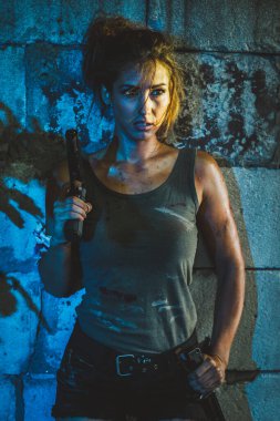 Tuğla duvarın önünde duran ve elinde silah tutan kolsuz tişörtlü çekici bir kadın..
