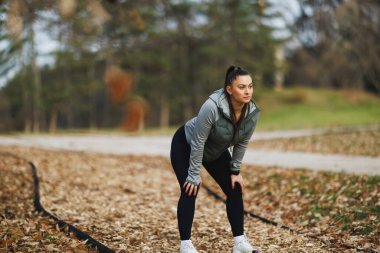 Genç bir kadın sonbaharda orman parkında yaprak kaplı bir patikada koşuya hazırlanıyor..